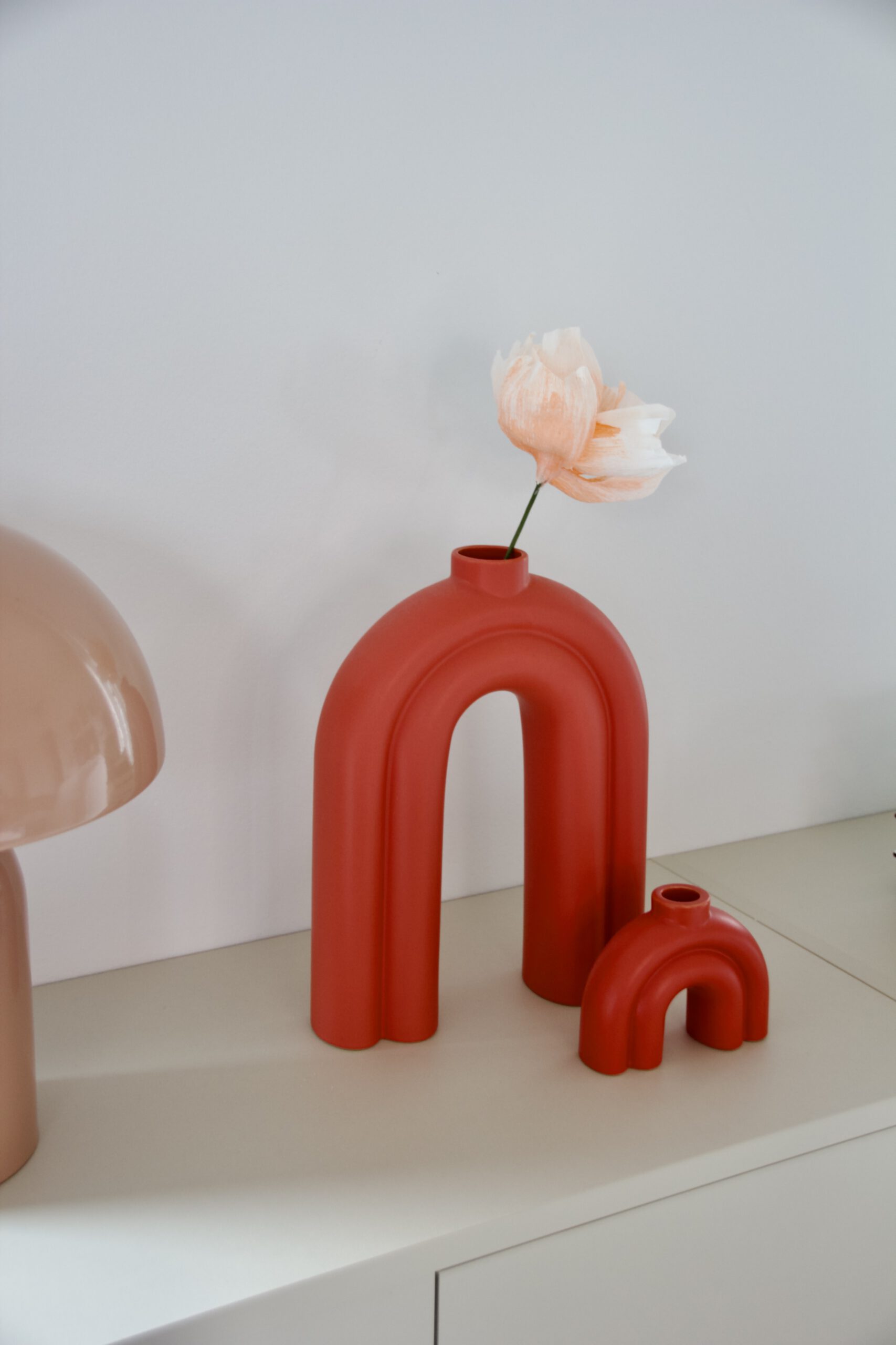 Zarte Papierblumen in blassem Orange in einer Coralroten Designer-Vase auf einem Regal